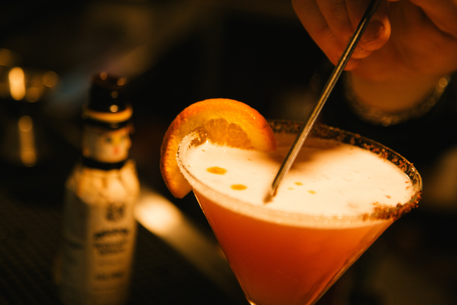 Un trago en copa con una rodaja de naranja en el borde, terminándose de preparar en la barra