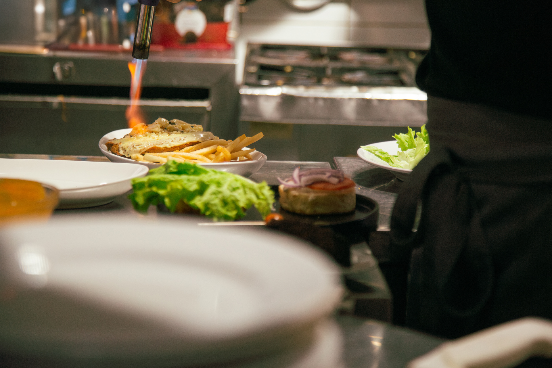 Platos listos en el pase de la cocina: una milanesa con papas, lechuga y una hamburguesa, con la llama de un soplete detrás